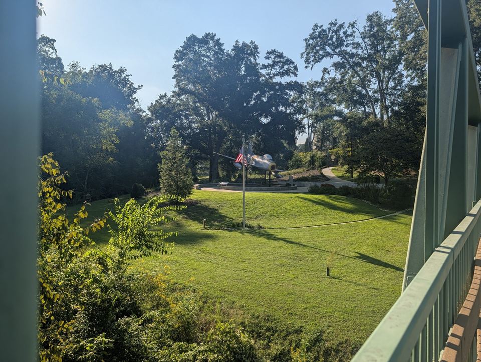 A sloping lawn and tall trees seen past a green bridge railing.