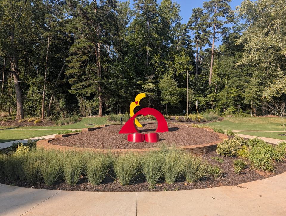 A red and yellow sculpture in a planted circle, surrounded by paths and trees.