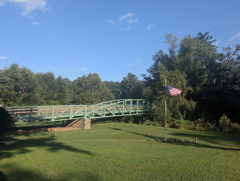 A green metal truss footbridge beyond a lawn, with an American flag on a pole.
